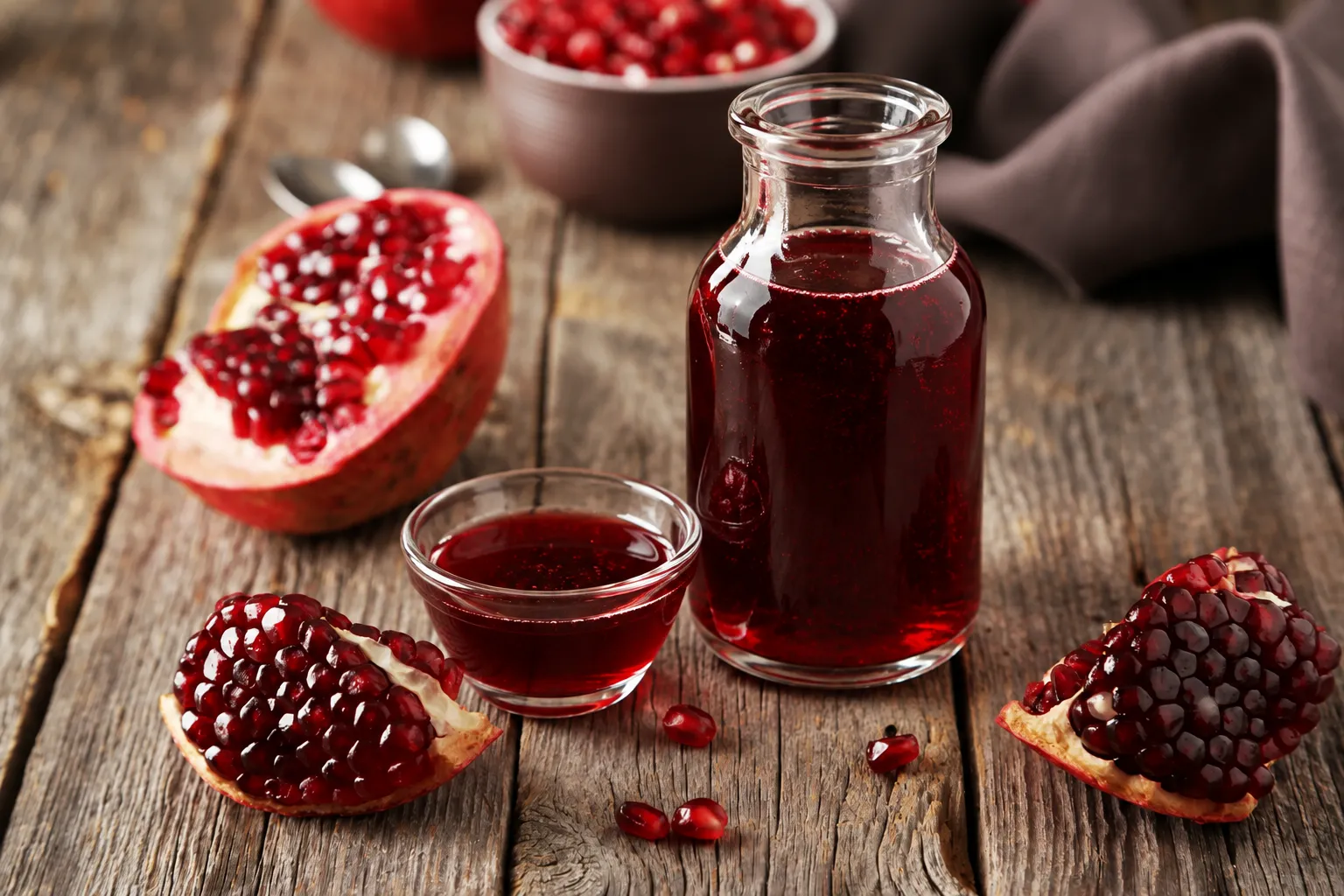 Glass bottle of deep red grenadine syrup beside a small glass and cut pomegranates on a wooden table.